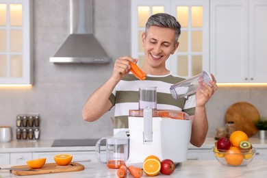 Photo of Smiling man putting fresh carrot into juicer at white marble table in kitchen