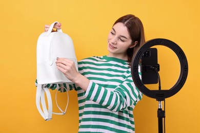 Photo of Fashion blogger reviewing backpack and recording video with smartphone and ring lamp on orange background