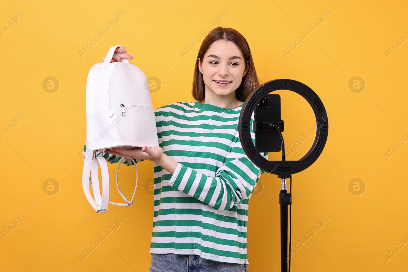 Photo of Fashion blogger reviewing backpack and recording video with smartphone and ring lamp on orange background