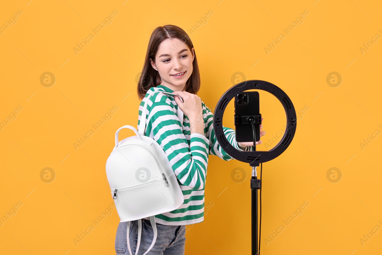 Photo of Fashion blogger reviewing backpack and recording video with smartphone and ring lamp on orange background
