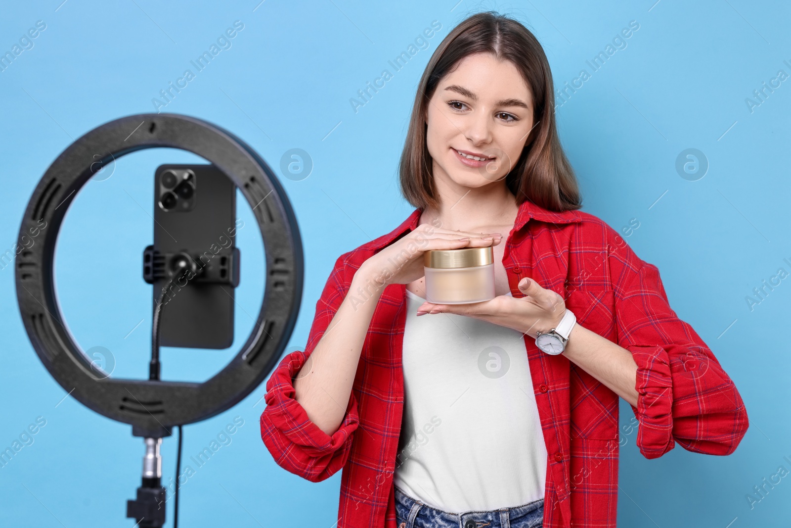 Photo of Beauty blogger reviewing cosmetic product and recording video with smartphone and ring lamp on light blue background