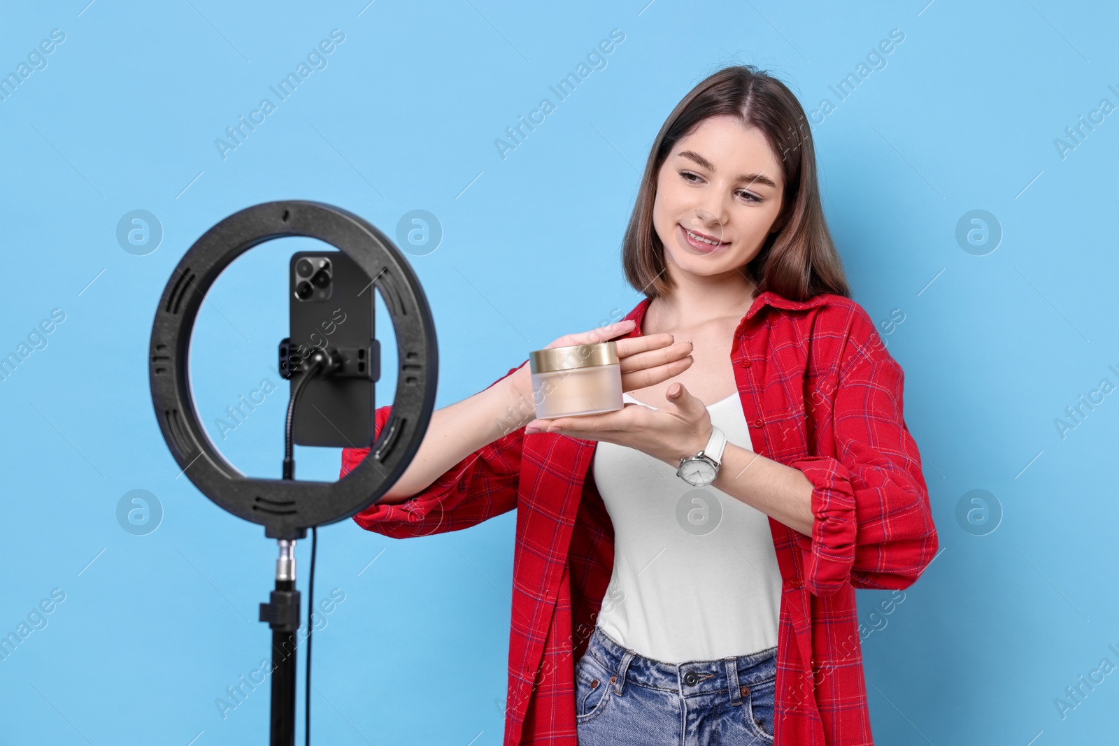Photo of Beauty blogger reviewing cosmetic product and recording video with smartphone and ring lamp on light blue background