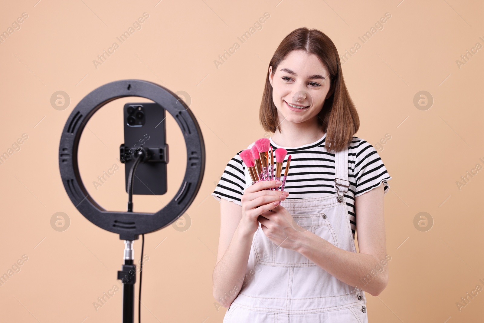 Photo of Beauty blogger reviewing makeup brushes and recording video with smartphone and ring lamp on beige background