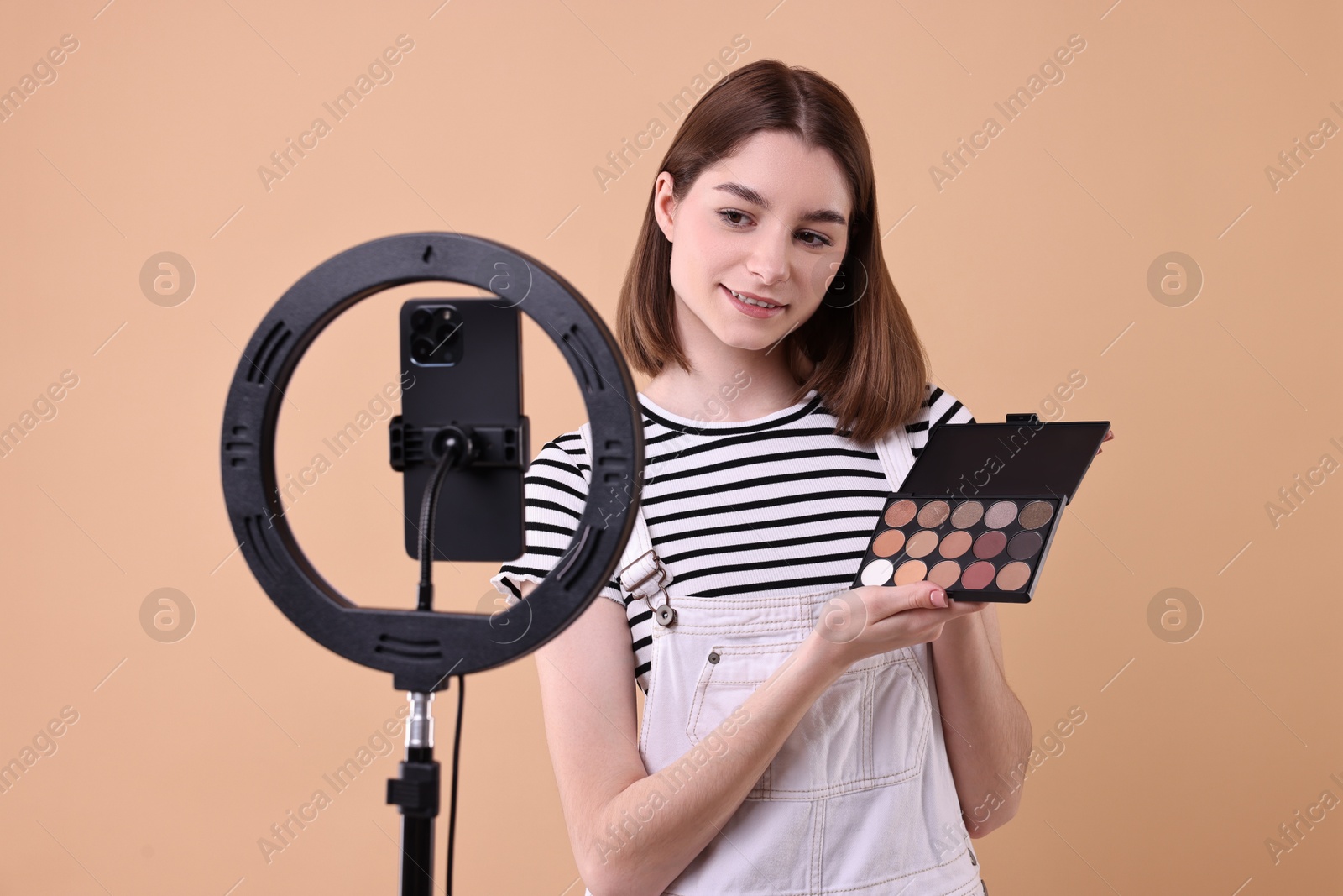 Photo of Beauty blogger reviewing eyeshadows and recording video with smartphone and ring lamp on beige background