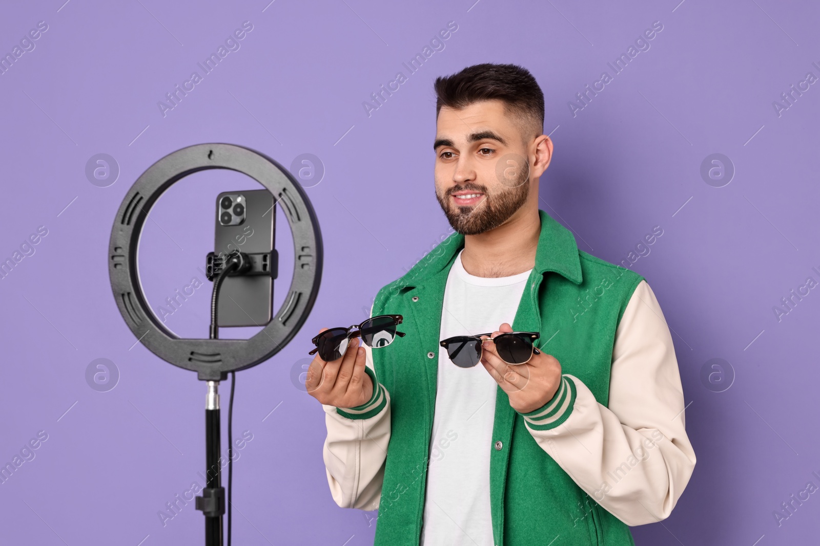 Photo of Fashion blogger reviewing sunglasses and recording video with smartphone and ring lamp on purple background