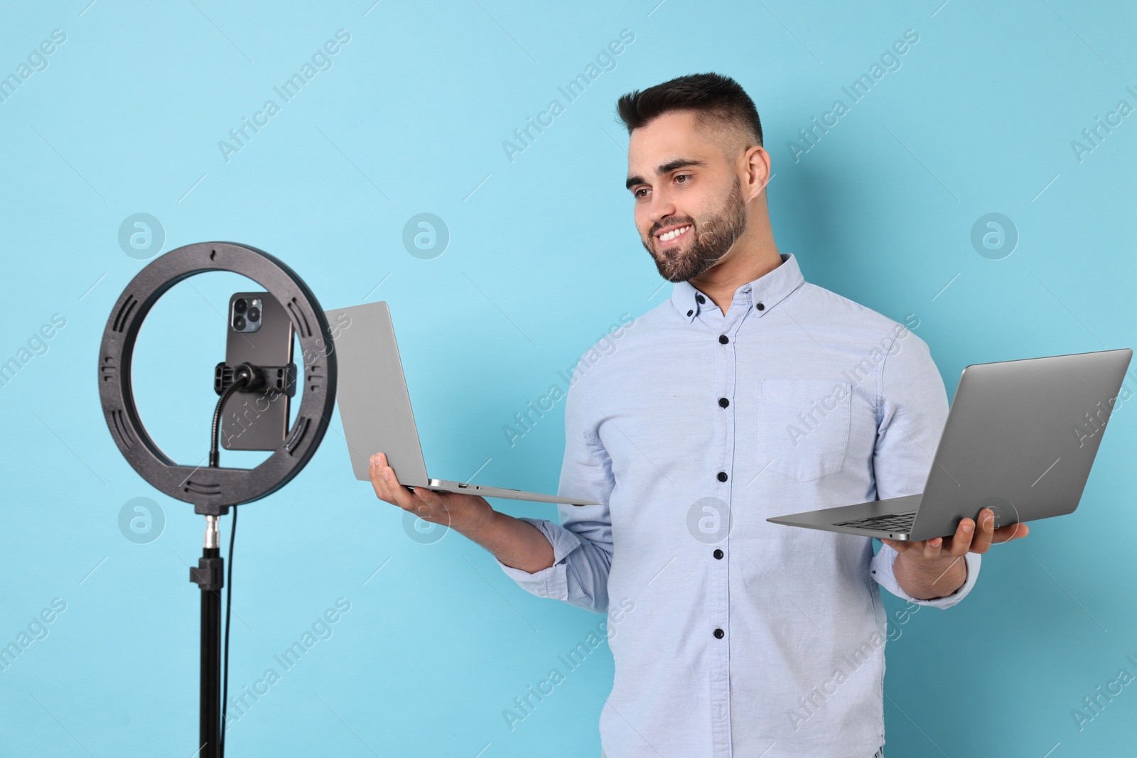 Photo of Technology blogger reviewing laptops and recording video with smartphone and ring lamp on light blue background