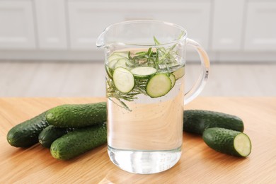 Refreshing cucumber water with rosemary in jug and vegetable on wooden table, closeup