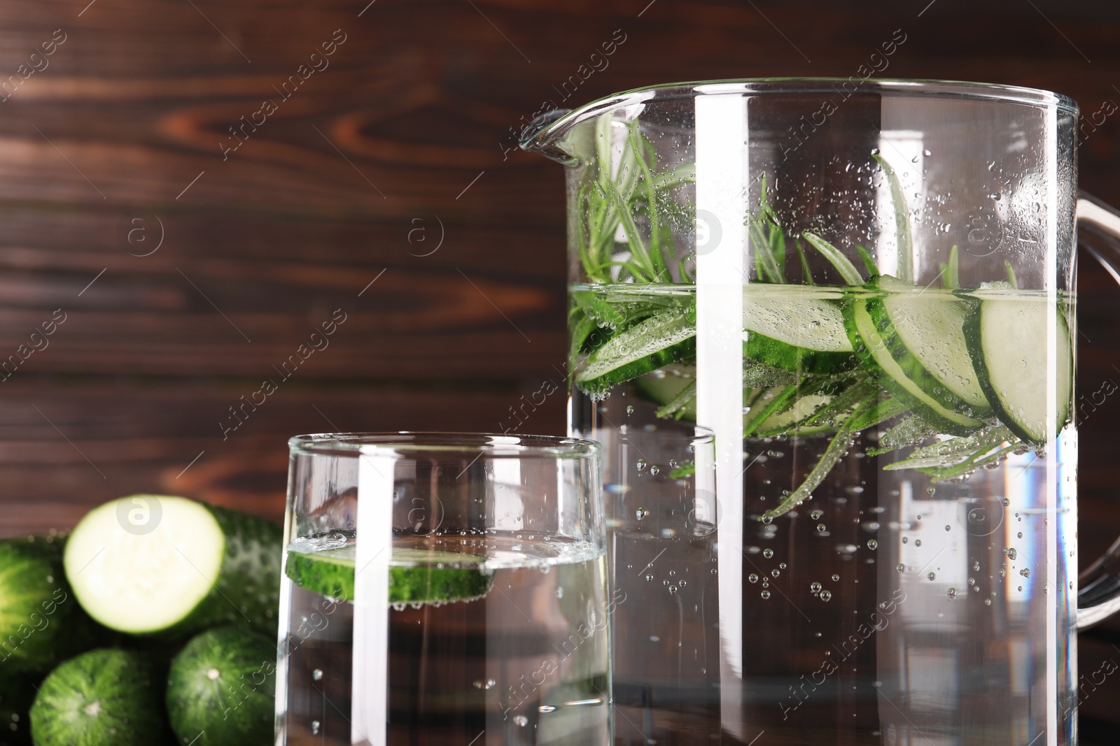 Photo of Refreshing cucumber water with rosemary in jug and glass against blurred background, closeup