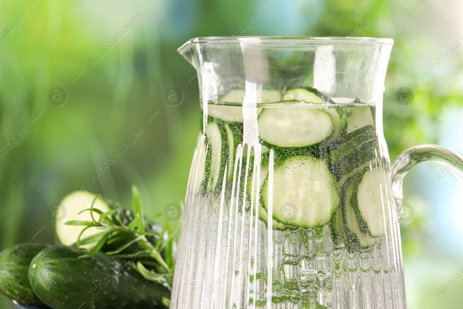 Photo of Refreshing cucumber water with rosemary in jug and vegetable against blurred green background, closeup