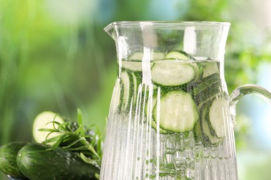 Photo of Refreshing cucumber water with rosemary in jug and vegetable against blurred green background, closeup