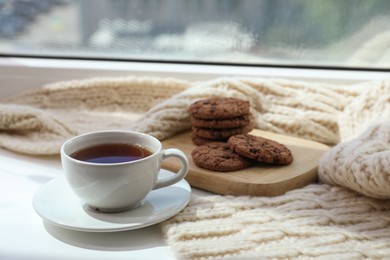 Photo of Beige knitted scarf, tea and cookies on windowsill, closeup