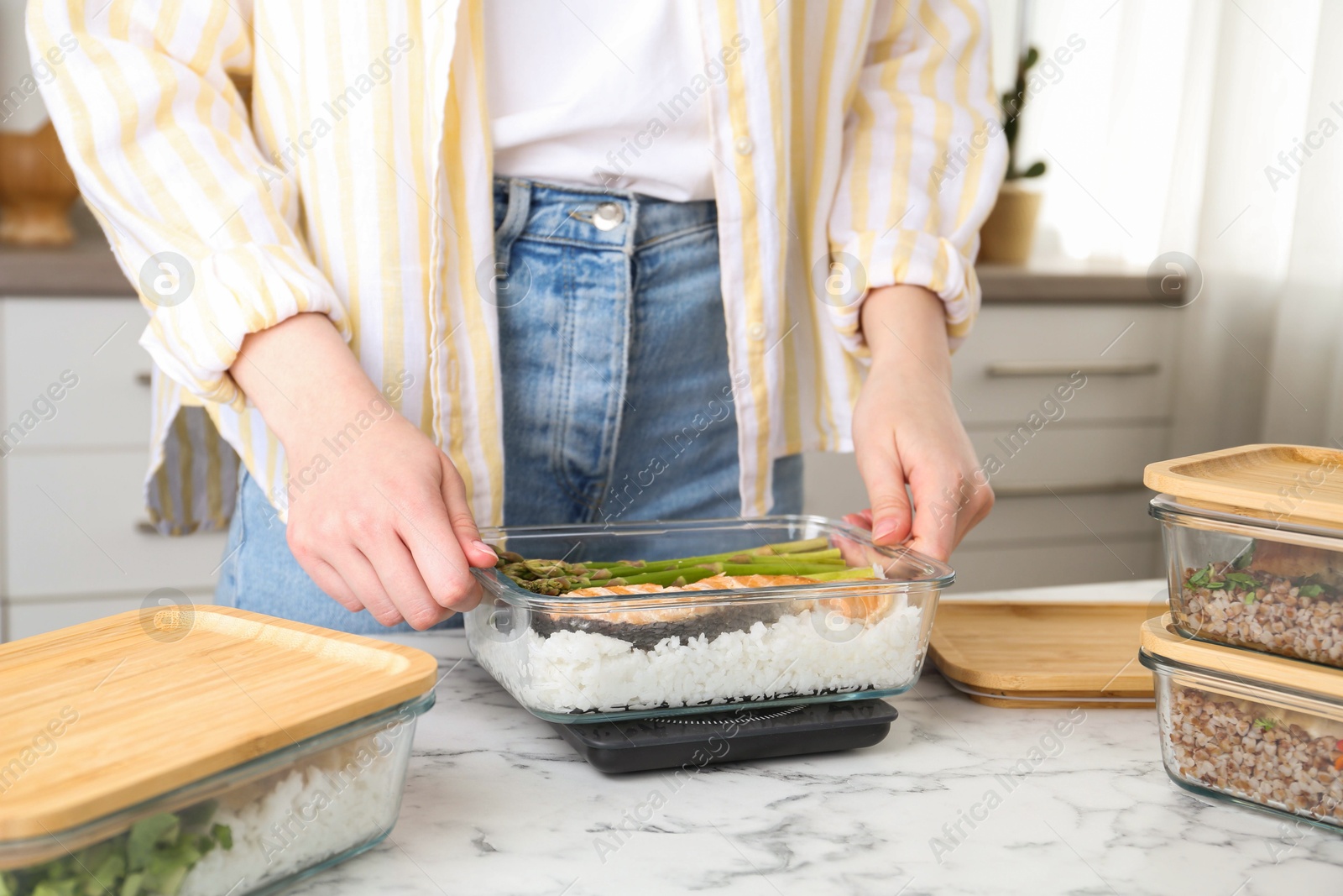 Photo of Healthy meal. Woman weighing glass container with food on kitchen scale at white marble table, closeup