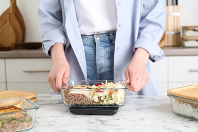 Healthy meal. Woman weighing glass container with food on kitchen scale at white marble table, closeup