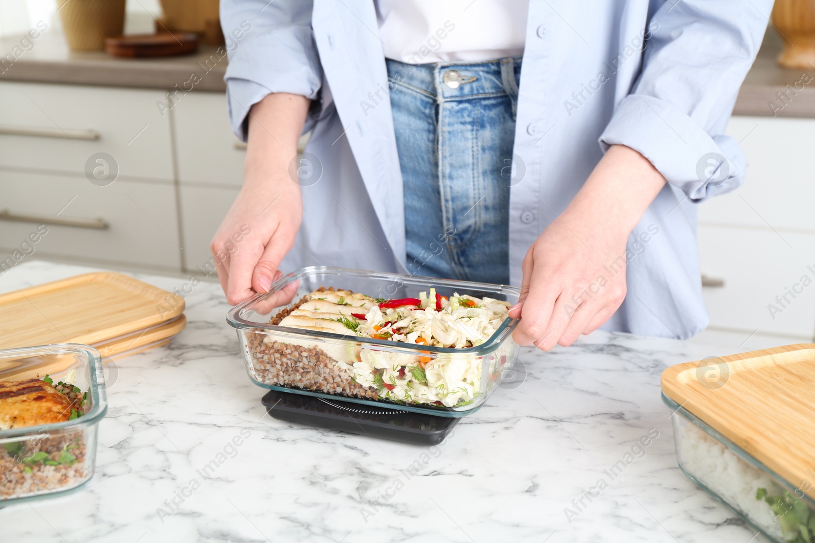 Photo of Healthy meal. Woman weighing glass container with food on kitchen scale at white marble table, closeup