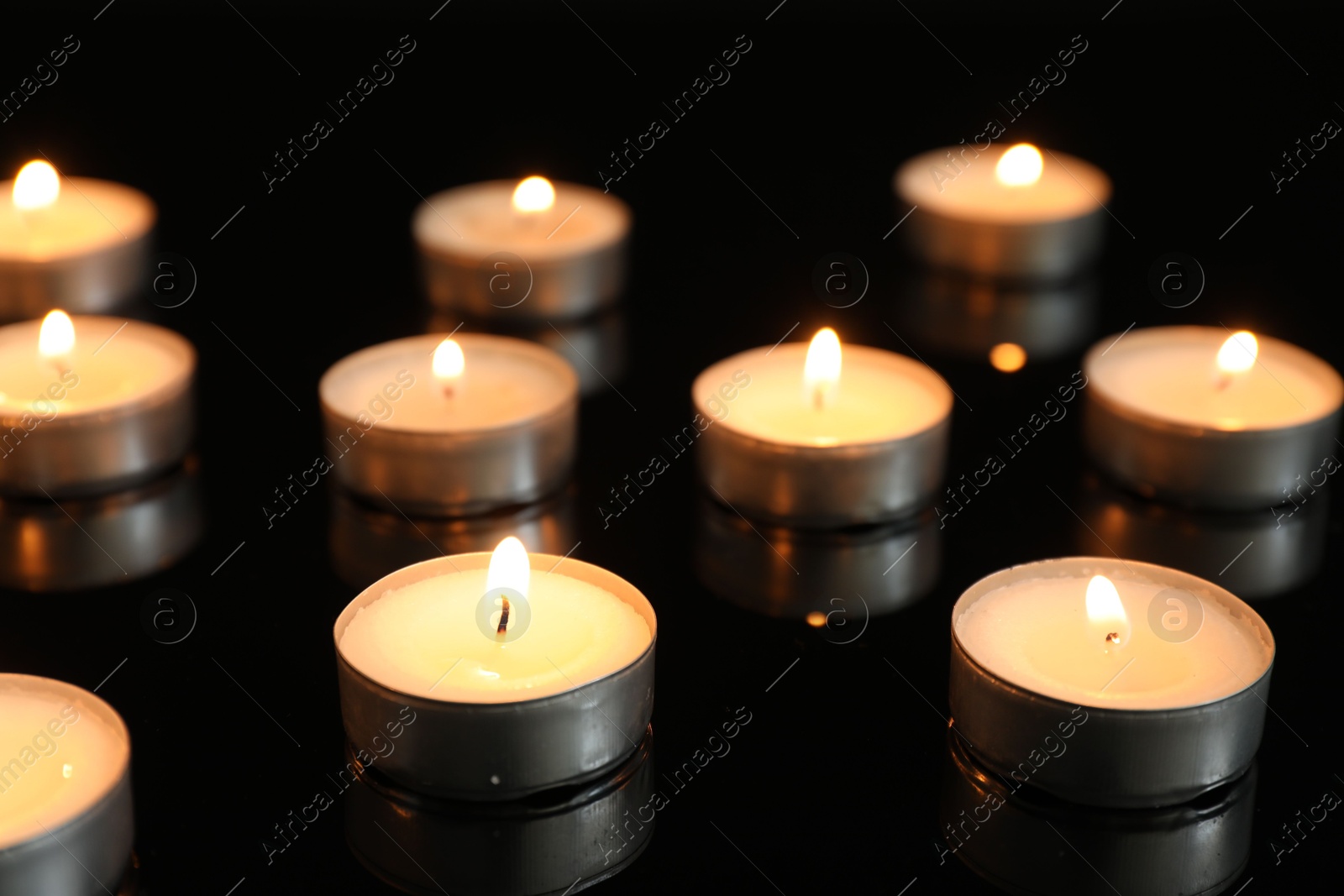 Photo of Many burning tealight candles on mirror surface against black background