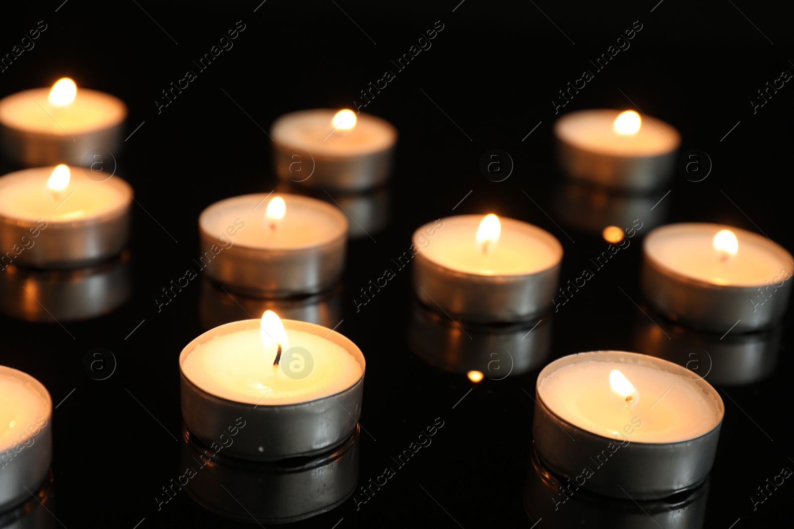 Photo of Many burning tealight candles on mirror surface against black background