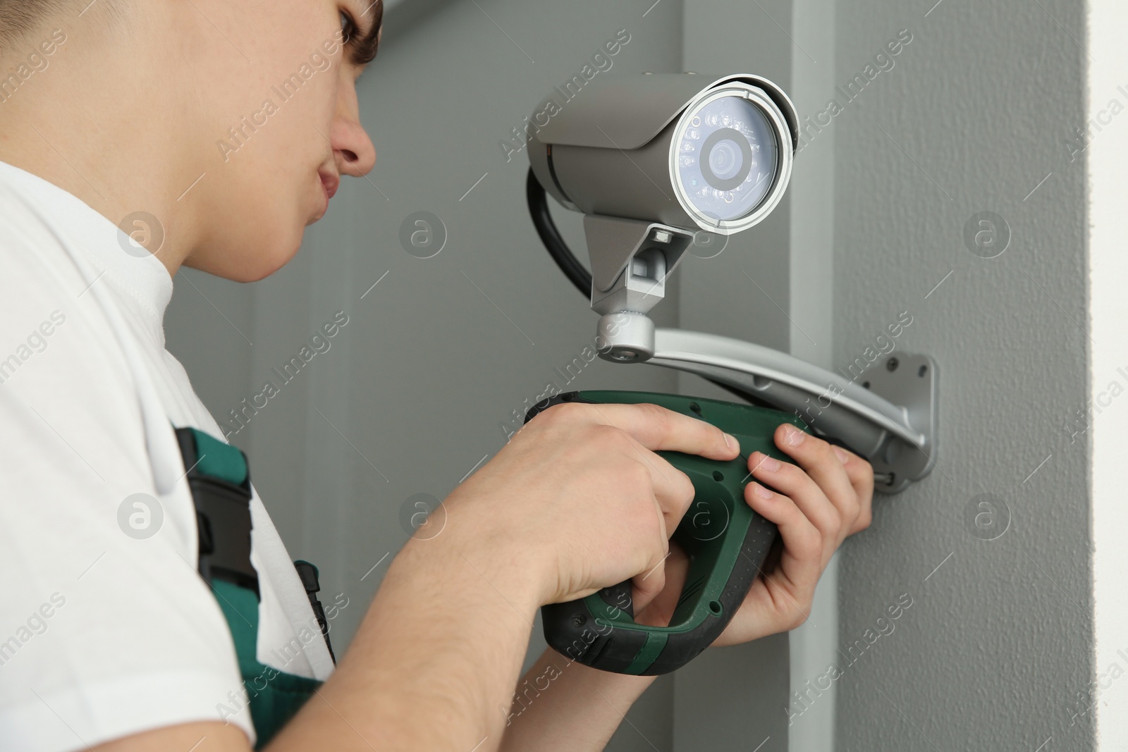 Photo of Technician with cordless electric screwdriver installing CCTV camera on wall indoors, closeup