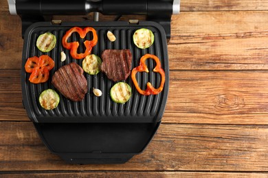 Photo of Electric grill with tasty meat steaks and vegetables on wooden table, top view. Space for text