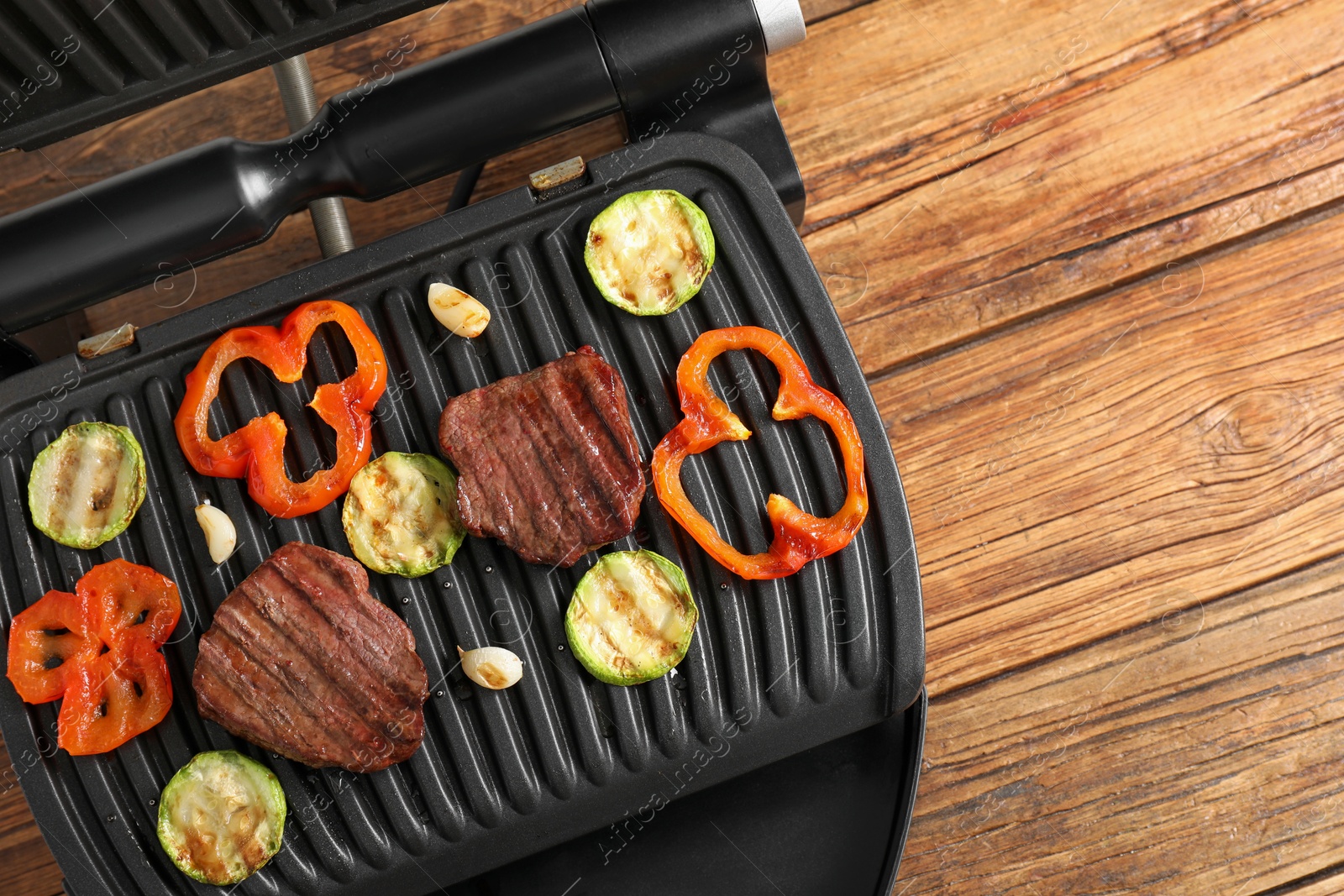 Photo of Electric grill with tasty meat steaks and vegetables on wooden table, top view. Space for text