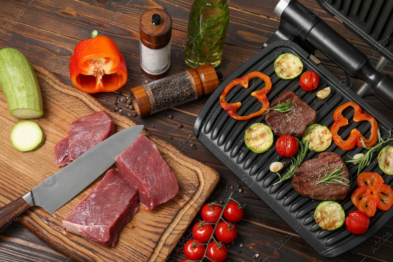Photo of Electric grill with meat, spices, knife and vegetables on wooden table, above view