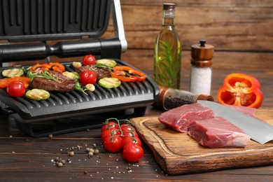 Electric grill with meat, spices, knife and vegetables on wooden table