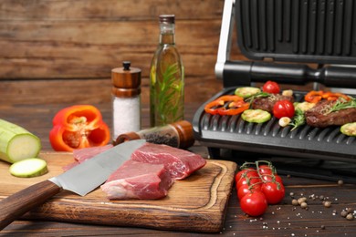 Photo of Electric grill with meat, spices, knife and vegetables on wooden table