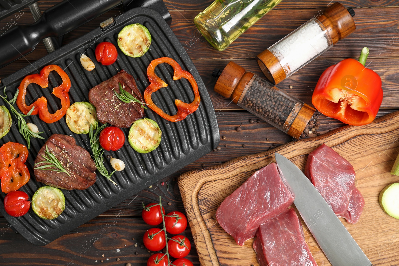 Photo of Electric grill with meat, spices, knife and vegetables on wooden table, flat lay