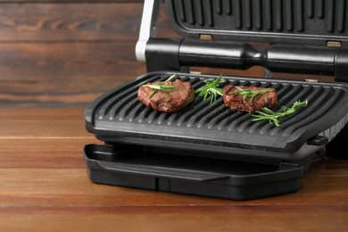 Photo of Electric grill with tasty meat steaks and rosemary on wooden table