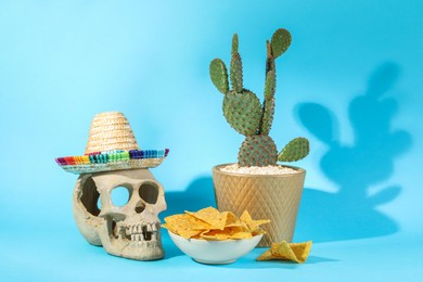 Human scull with Mexican sombrero hat, cactus and nachos chips on light blue background