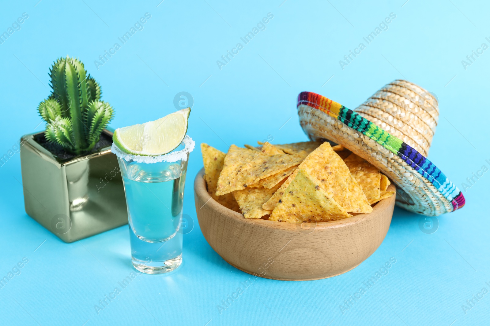 Photo of Mexican sombrero hat, cactus, nachos chips and tequila on light blue background