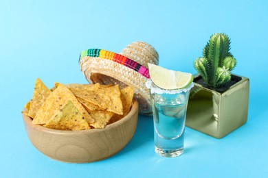 Photo of Mexican sombrero hat, cactus, nachos chips and tequila on light blue background