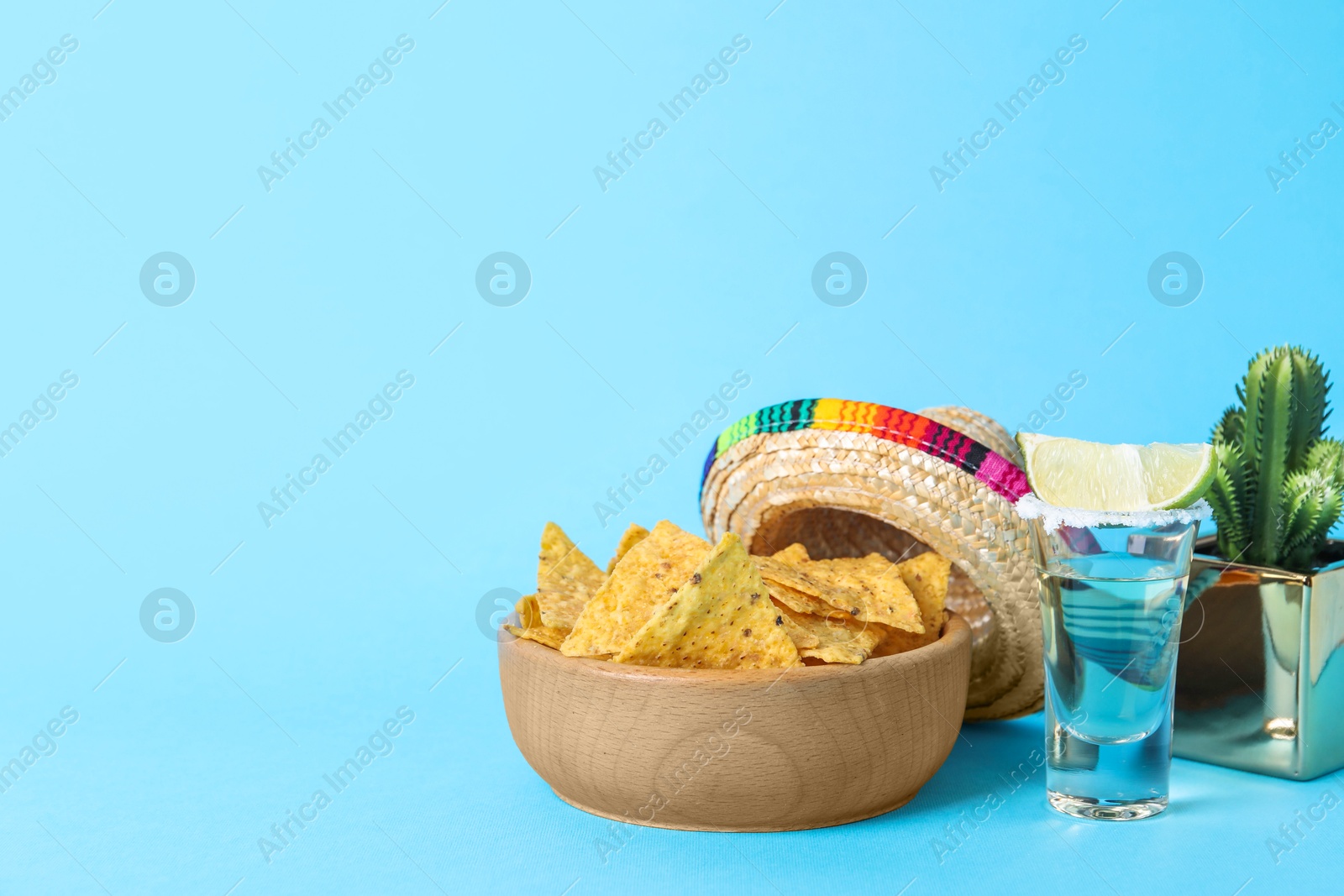Photo of Mexican sombrero hat, cactus, nachos chips and tequila on light blue background. Space for text