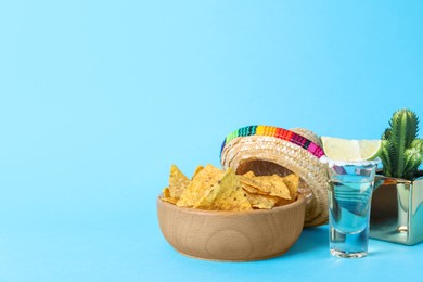 Photo of Mexican sombrero hat, cactus, nachos chips and tequila on light blue background. Space for text
