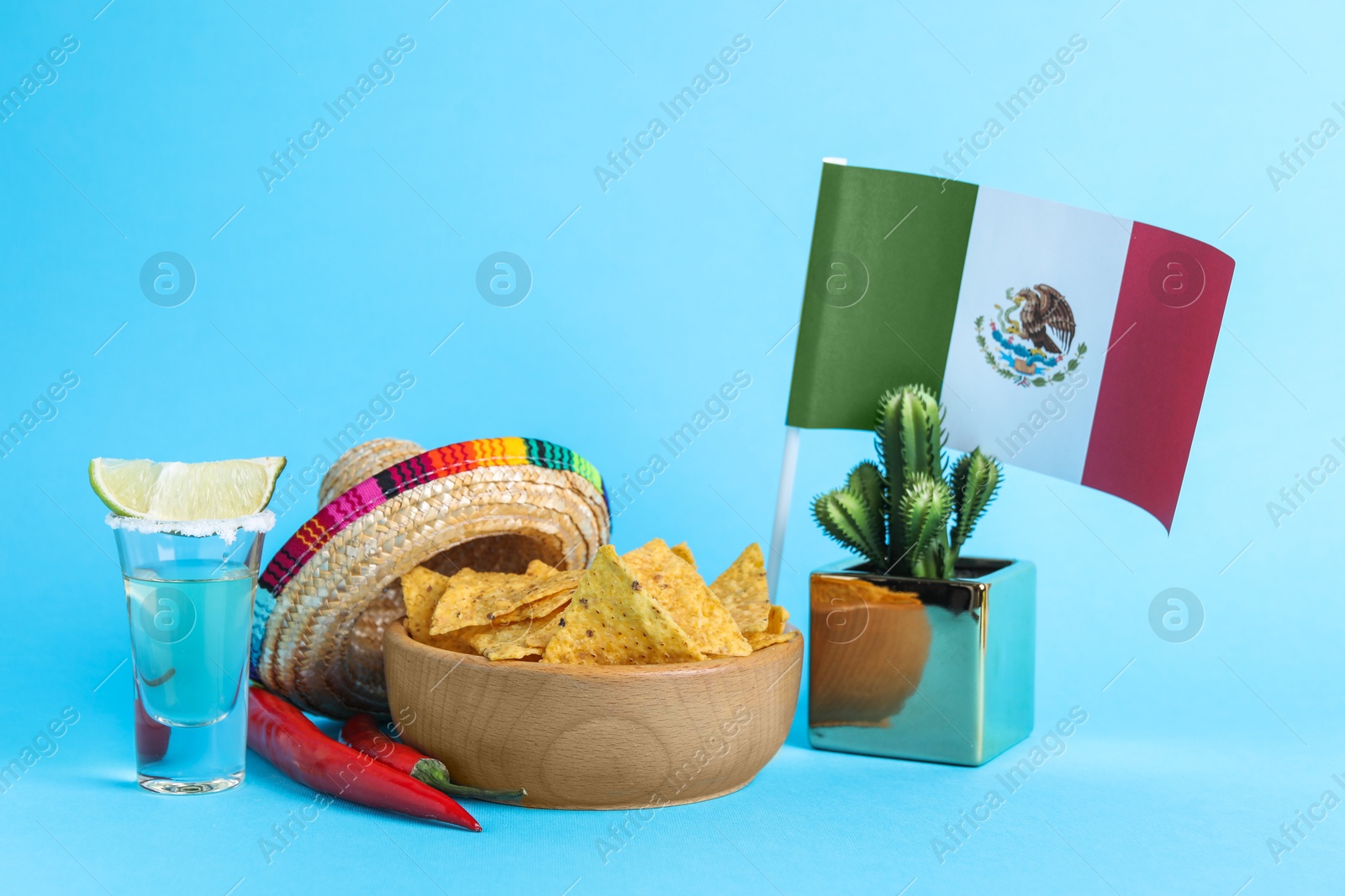 Photo of Mexican flag, sombrero hat, chili peppers, nachos chips and tequila on light blue background