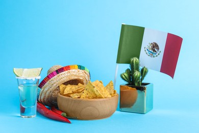 Photo of Mexican flag, sombrero hat, chili peppers, nachos chips and tequila on light blue background