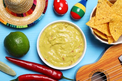 Delicious guacamole with nachos chips, Mexican sombrero hat, ukulele and maracas on light blue wooden table, flat lay