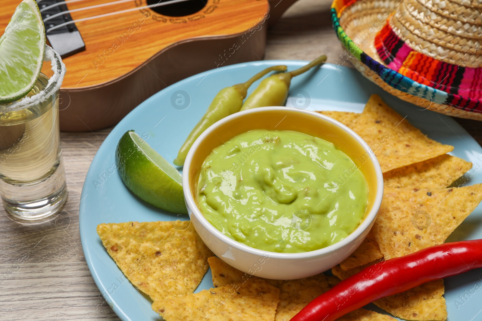 Photo of Guacamole, nachos chips, tequila, Mexican sombrero hat and ukulele on wooden table