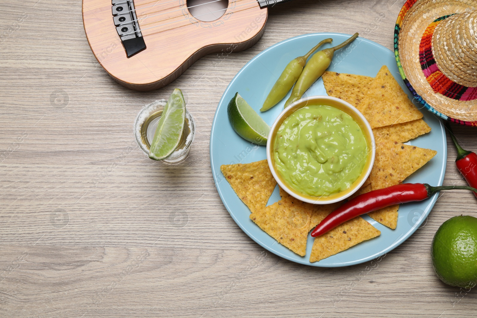 Photo of Guacamole, nachos chips, tequila, Mexican sombrero hat and ukulele on wooden table, flat lay. Space for text