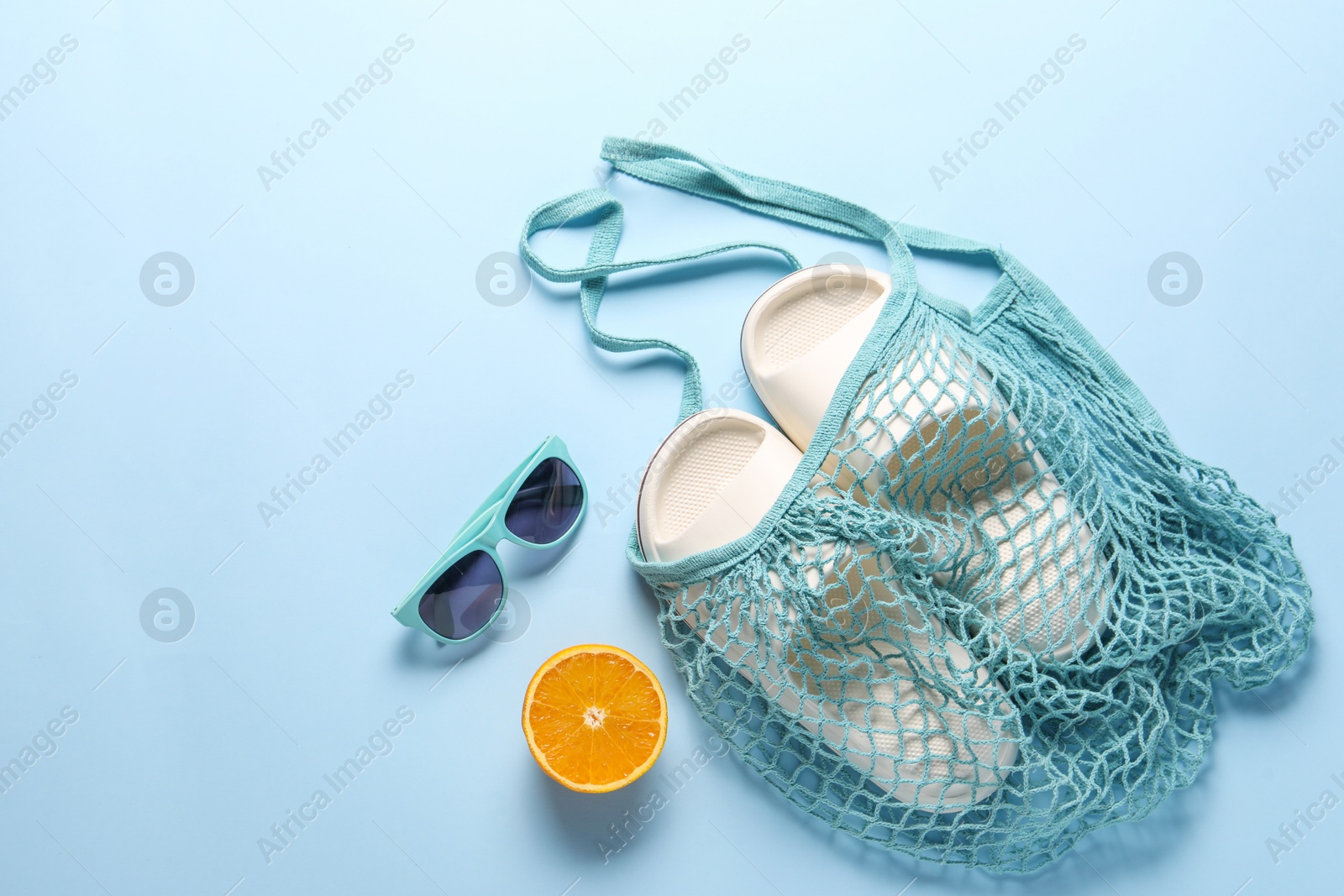 Photo of String bag, sunglasses, orange and slippers on light blue background, top view. Space for text