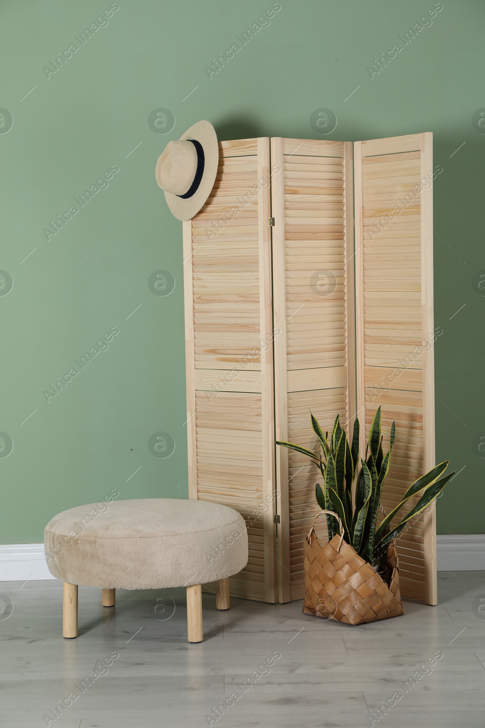 Photo of Wooden folding screen, pouffe, bag, houseplant and hat near green wall indoors