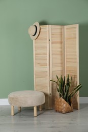 Wooden folding screen, pouffe, bag, houseplant and hat near green wall indoors