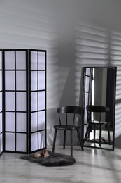 Photo of Folding screen, shoes, chair and mirror near white wall indoors
