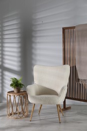 Wooden folding screen, blanket, armchair, side table and houseplant near light wall indoors