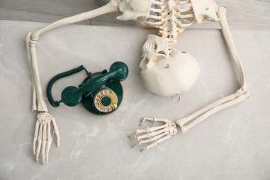 Photo of Waiting concept. Human skeleton at grey marble table with corded telephone, top view