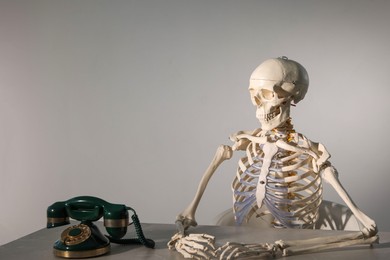 Photo of Waiting concept. Human skeleton at table with corded telephone against grey background