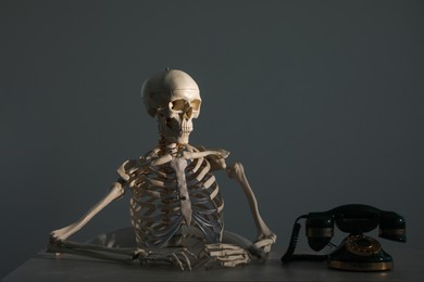 Waiting concept. Human skeleton at table with corded telephone against grey background