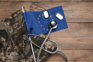 Photo of Stethoscope, flag of European Union, tags and military uniform on wooden table, flat lay. Space for text