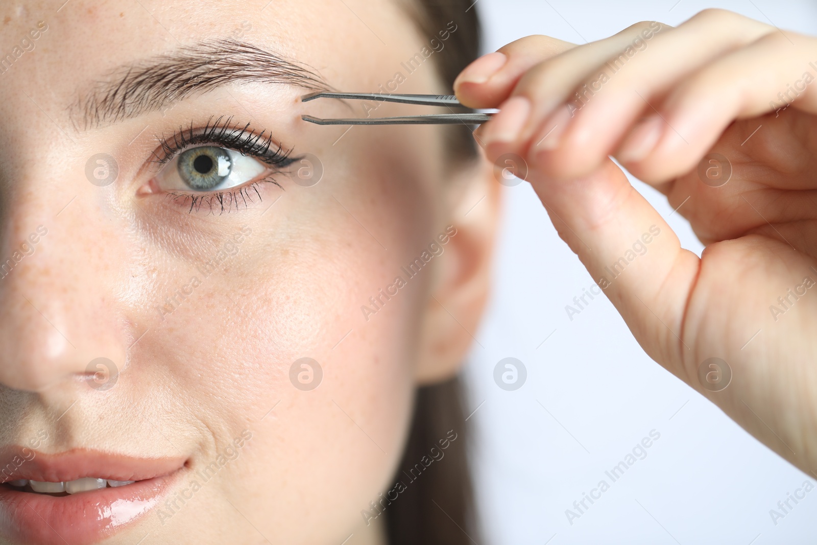 Photo of Young woman plucking eyebrow with tweezers on light background, closeup