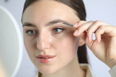 Young woman plucking eyebrow with tweezers on light background, closeup