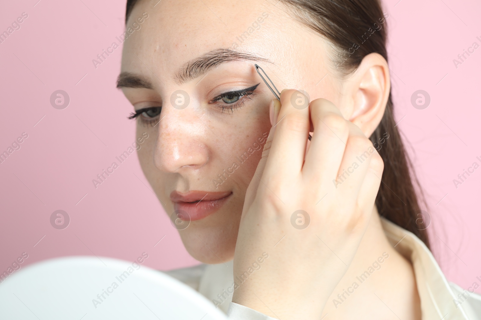 Photo of Young woman plucking eyebrow with tweezers on pink background, closeup